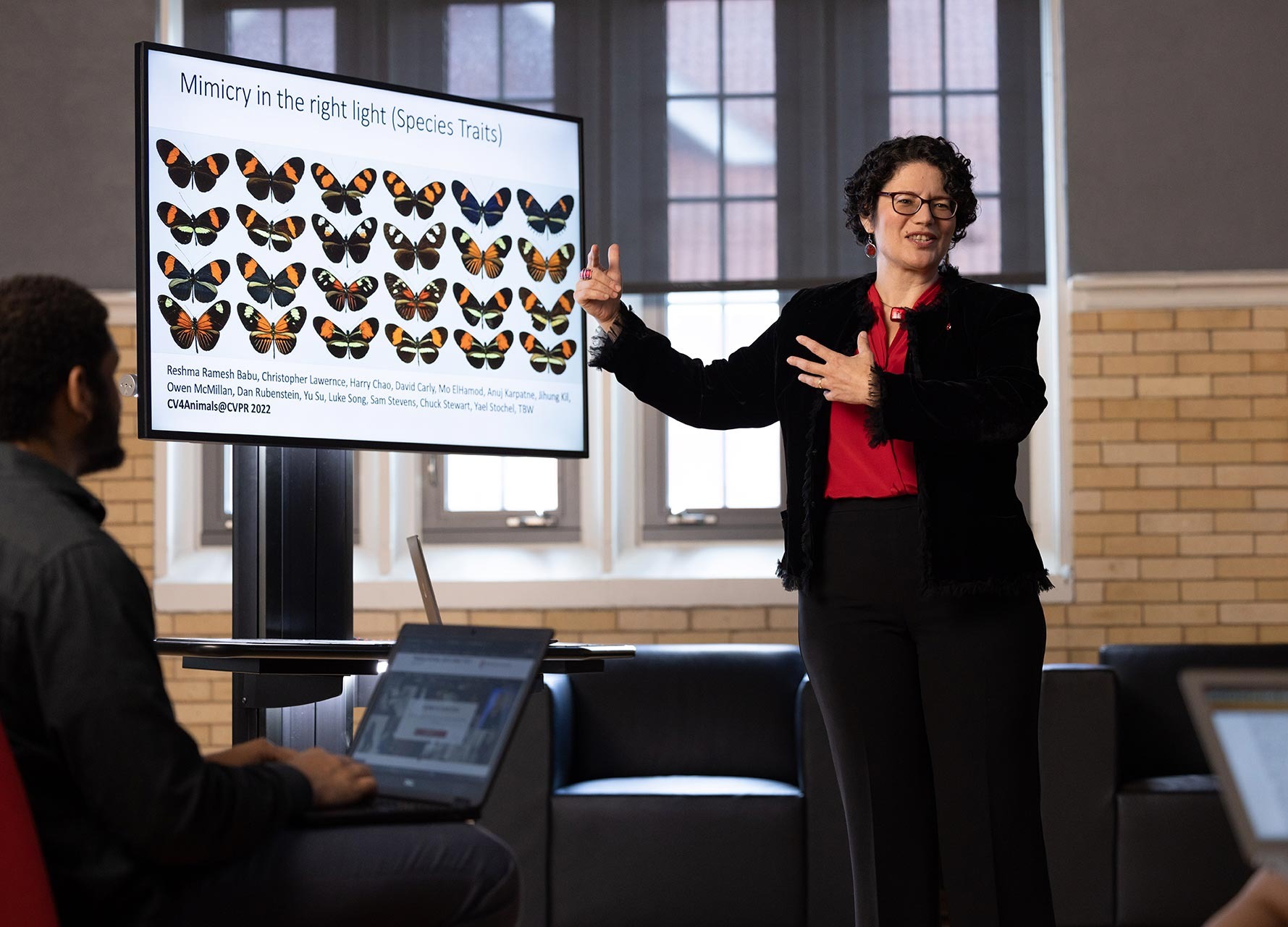A presenter in a black blazer and red shirt gestures toward a screen displaying rows of butterflies.