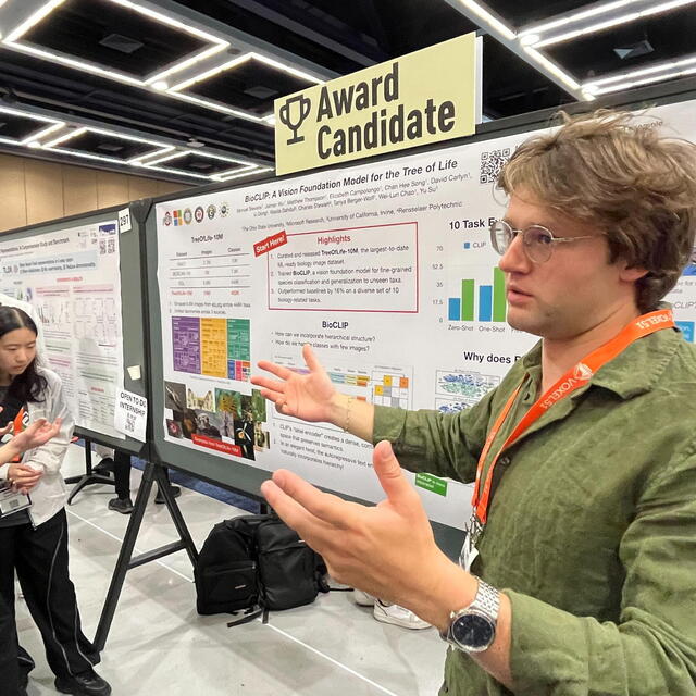 A researcher wearing a green shirt and glasses gestures toward a scientific poster marked with a yellow "Award Candidate" sign.