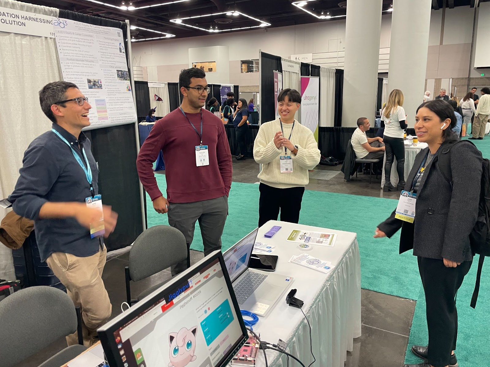Four people talk and laugh at a conference booth with laptops and a pink character on a monitor.