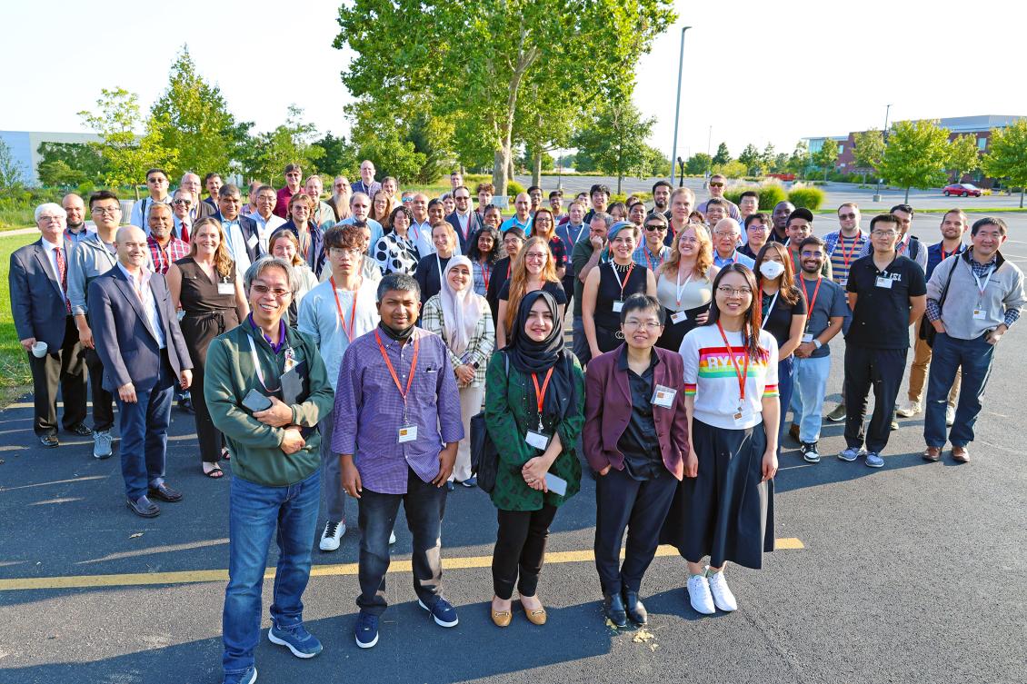 A large group of people stands outside on a paved area for a photo. They are wearing lanyards and smiling under a clear sky with green trees.