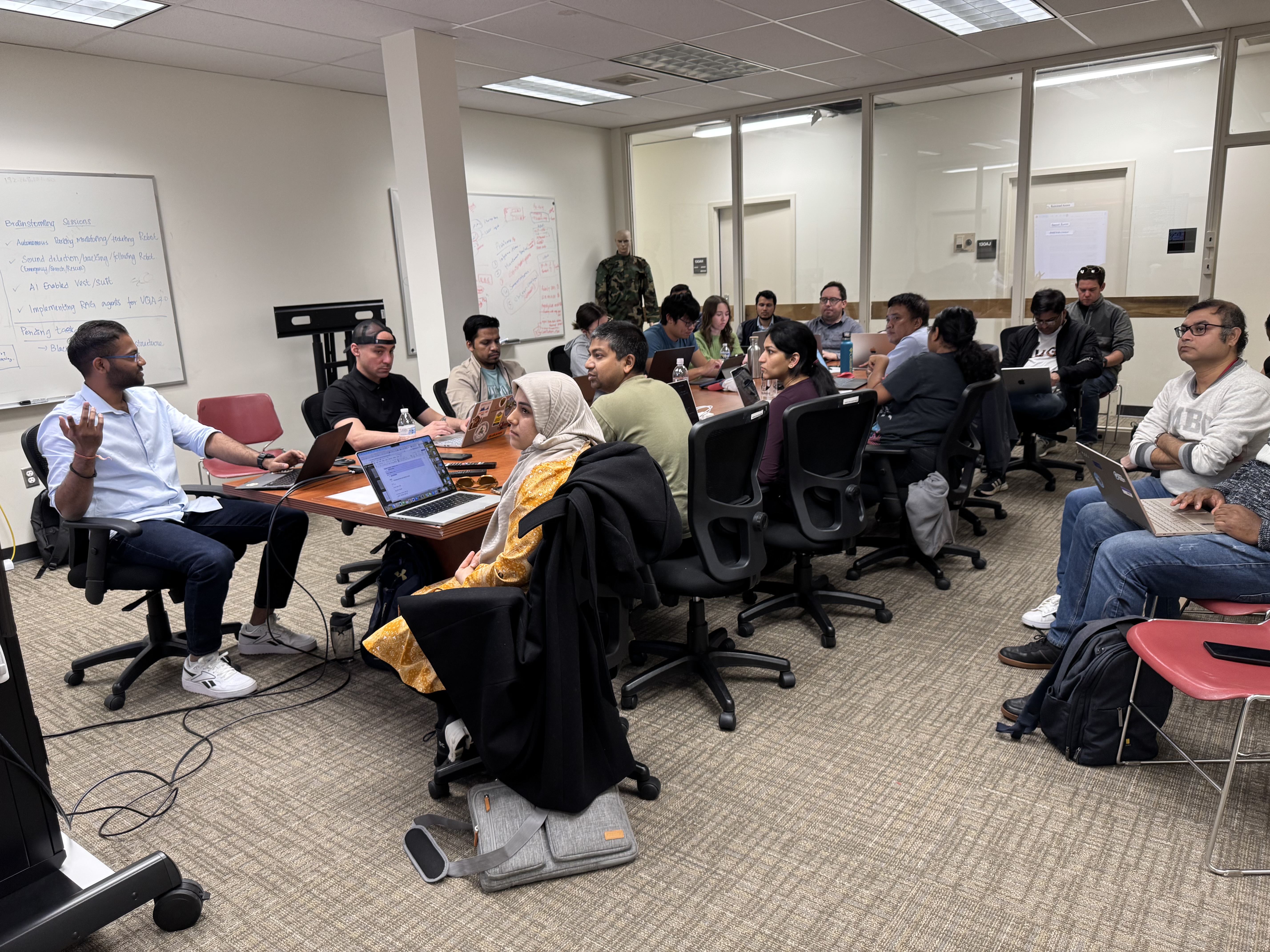A large group of students and professionals sit around a long conference table in a collaborative workshop setting.