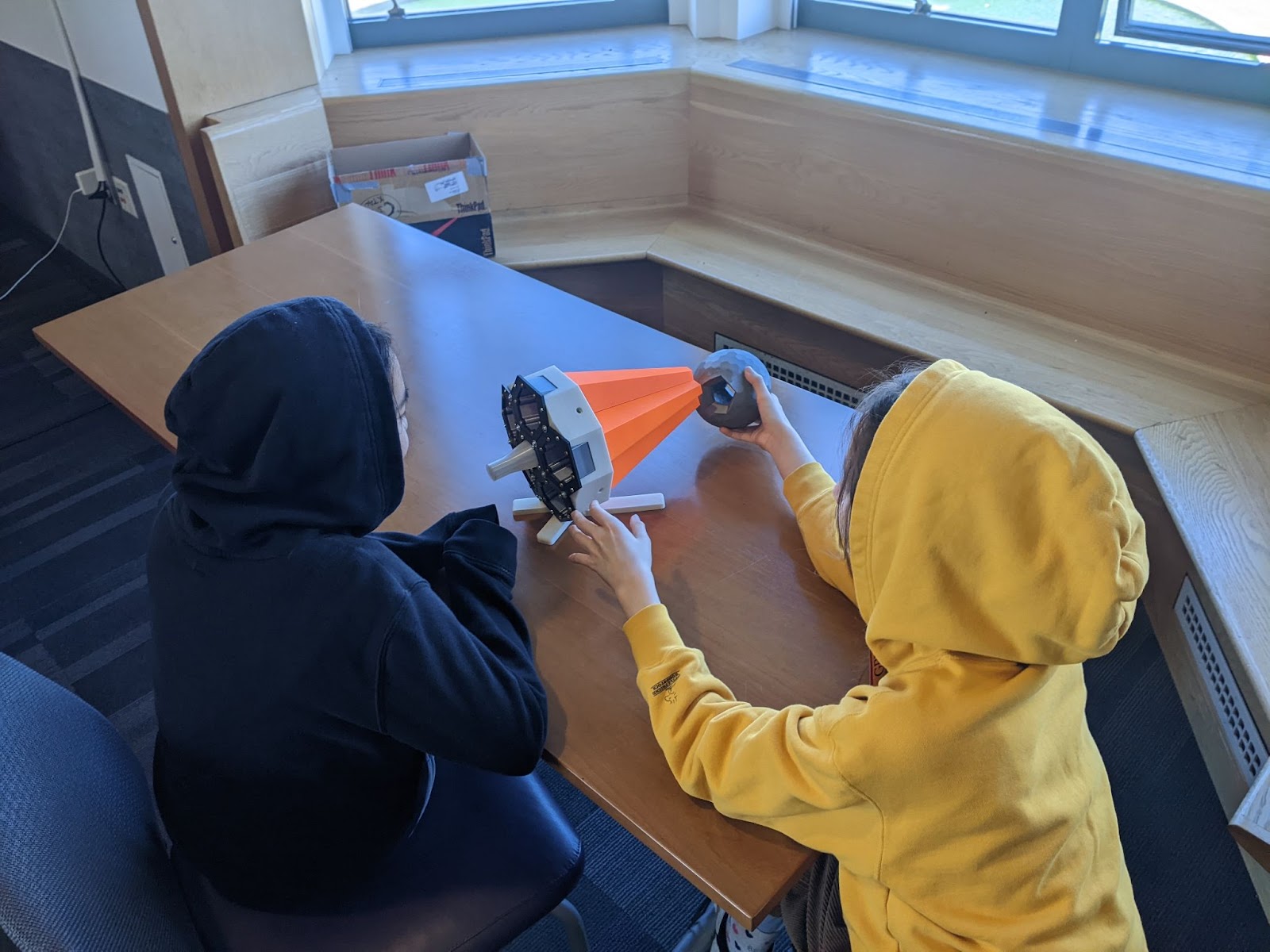 Two kids in hoodies sit at a table, interacting with a 3D-printed conical orange and white scientific model and a grey sphere.